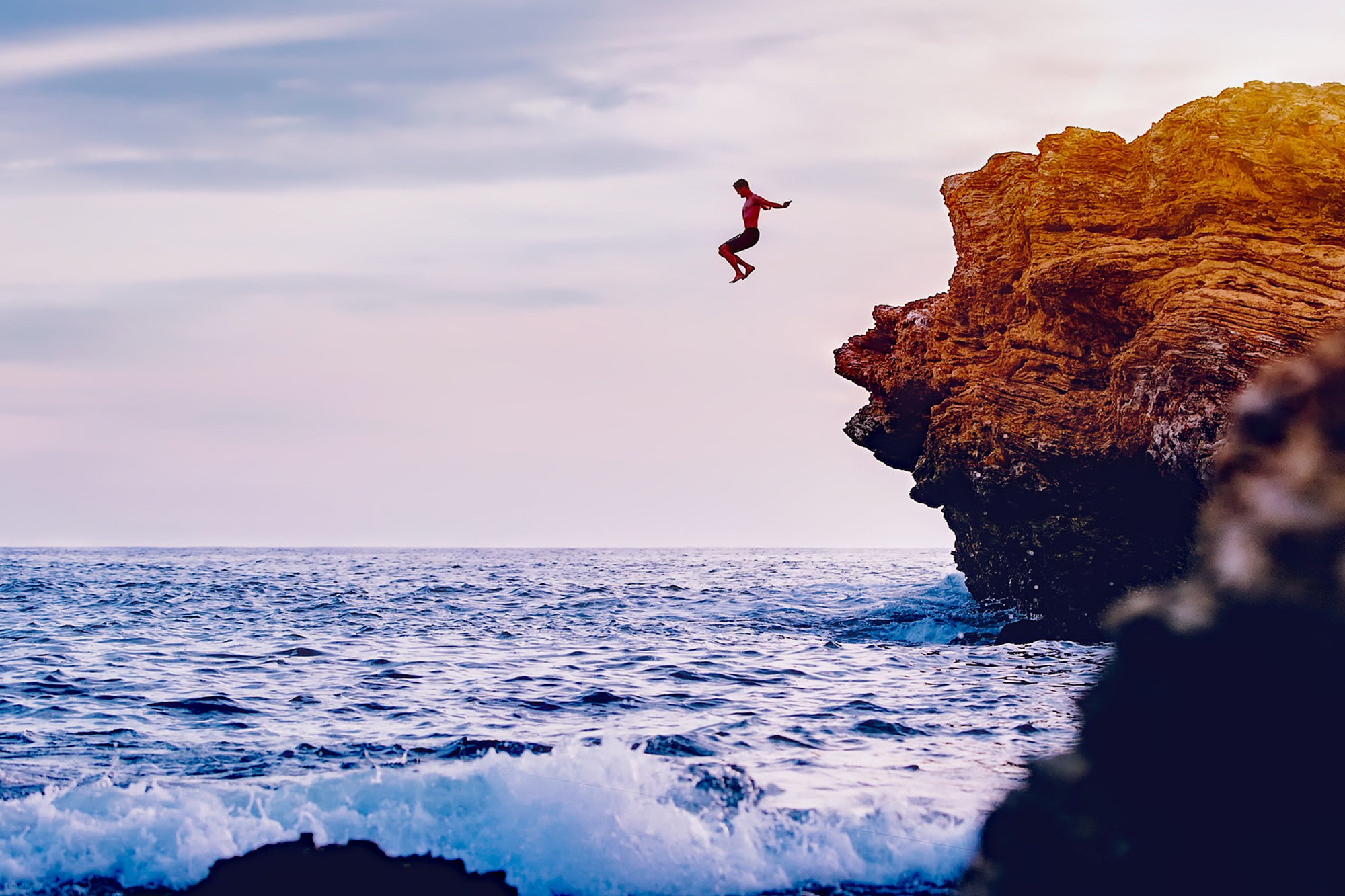 Man jumping off rocks into the sea with sunset